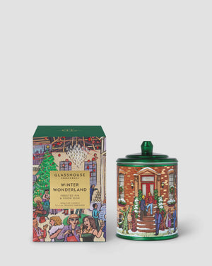 A Christmas-themed scented candle and its matching box from the brand GLASSHOUSE PARADISE, featuring a colorful winter village illustration on both the candle container and the packaging.