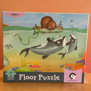 Puzzle - Māui Dolphin Mischief - 24 piece A boxed children's floor puzzle by Lakes Pieces featuring an underwater illustration of two dolphins, a brown rodent on a log, and colorful coral.