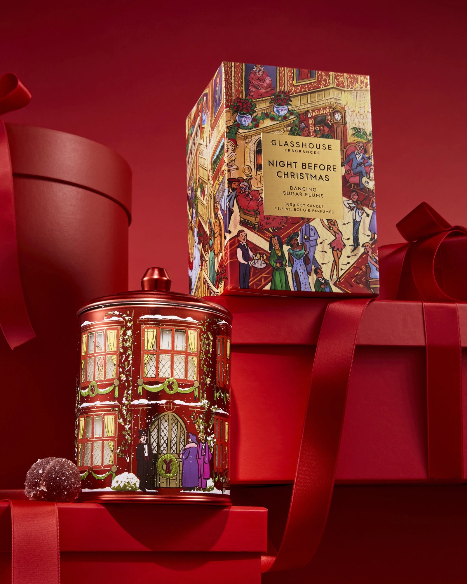 A Christmas-themed glass jar candle and its matching retail box by Glasshouse, featuring a festive red building illustration with holiday figures.