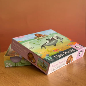 A children's floor puzzle box shown on a wooden tabletop with a warm terracotta background. The box features colorful cartoon animal illustrations, including a beaver, a porcupine, and a penguin, with text reading 'Kooi Floor Puzzle' and 'Floor Puzzle' and '24 LARGE PIECES'.