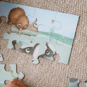 A children's jigsaw puzzle featuring a brown badger and a bottle-neck turtle on a beach, shown partially assembled on a beige woven rug with a child's hand visible at the bottom.