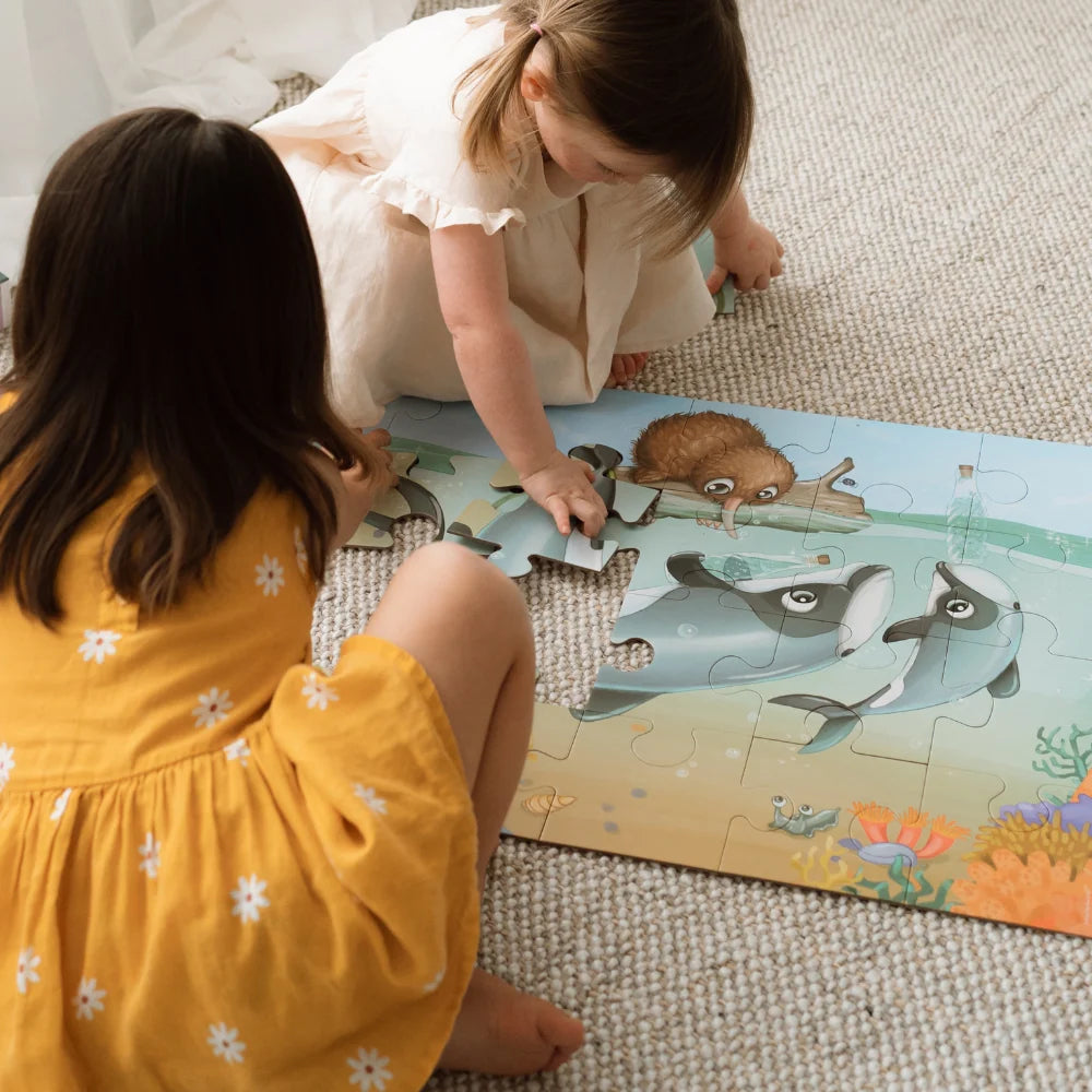 Puzzle - Māui Dolphin Mischief - 24 piece A boxed children's floor puzzle by Lakes Pieces featuring an underwater illustration of two dolphins, a brown rodent on a log, and colorful coral.