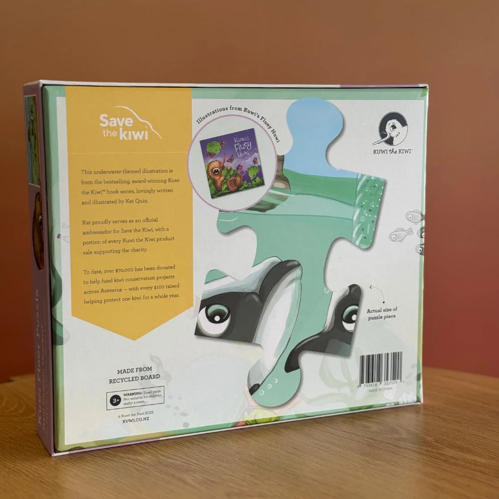 The retail packaging box for the 'Save the kiwi' Kiwi Kiin' puzzle kit, standing on a wooden surface against a warm brown background. The box features a large green kiwi puzzle piece illustration with cartoon eyes and a black-and-white kiwi graphic, alongside product details and a barcode.