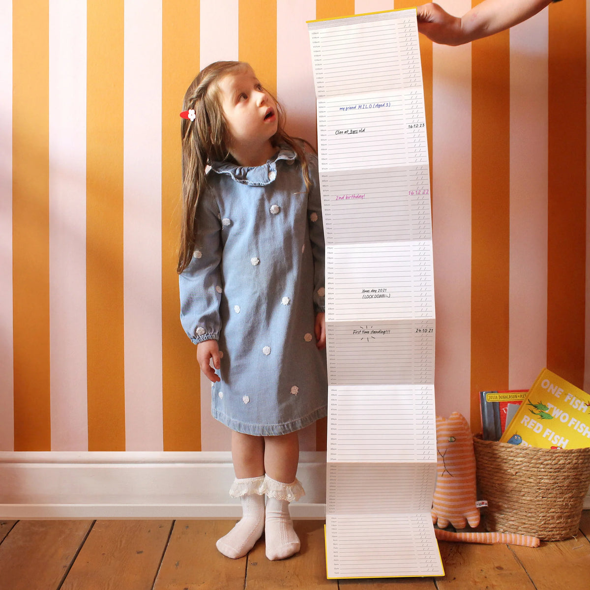 Suck UK My Height Chart A children's collapsible height chart book shown standing upright on a light gray background. The cover is white with bold yellow text reading 'MY HEIGHT CHART,' plus illustrations of folded pages and a young girl in a light blue dress standing beside the chart area.