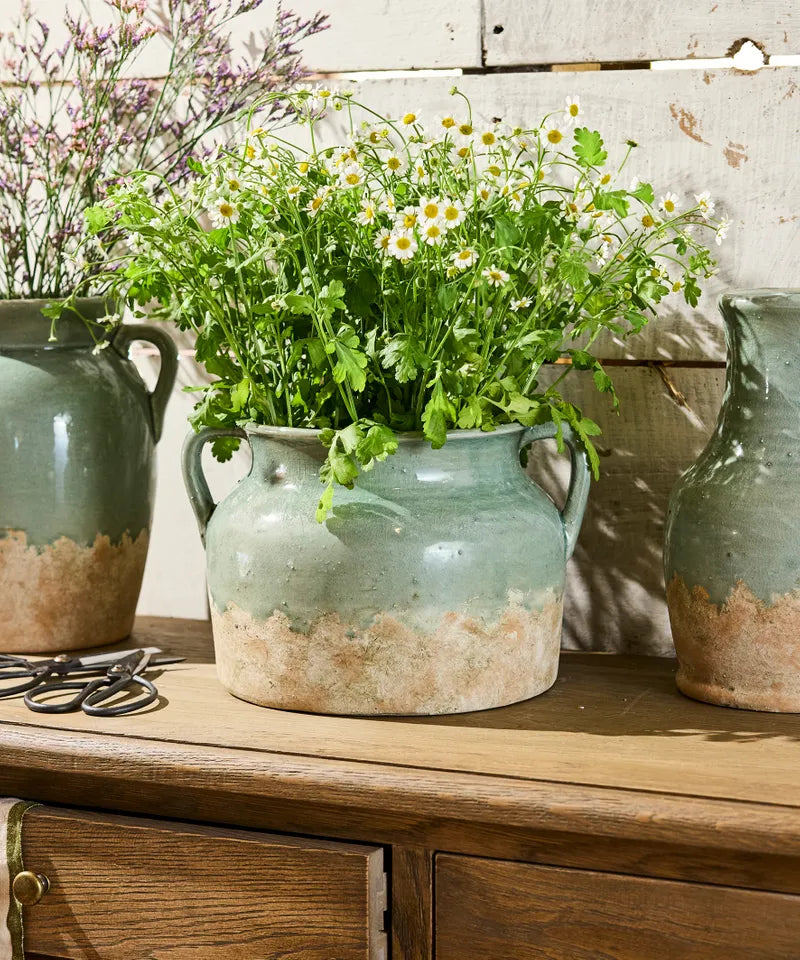 French Country Collections Varese Vessel Wide Pale Blue A rustic ceramic vase with two side handles, featuring a textured glaze that transitions from a muted sage green at the top to a distressed, earthy beige at the bottom rustic ceramic pots