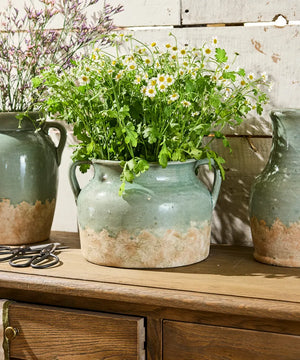 French Country Collections Varese Vessel Wide Pale Blue A rustic ceramic vase with two side handles, featuring a textured glaze that transitions from a muted sage green at the top to a distressed, earthy beige at the bottom rustic ceramic pots